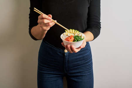 female hands holding chopsticks poke bowl with salmon, avocado rice, Chuka Salad,sweet onions, quail eggs sprinkled with white and black sesame on white backgroundの写真素材