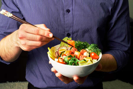 man in a shirt holds poke bowl with salmon, avocado, cucumbers, arugula, broccoli, rice, carrots,...