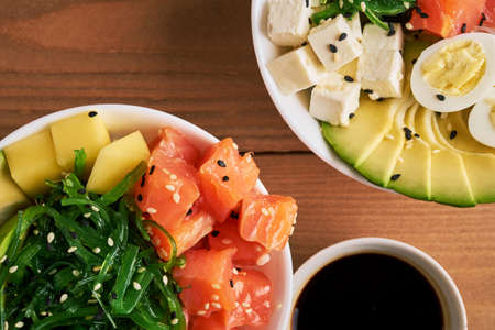 Raw Organic Poke Bowl with with rice, avocado, salmon, mango, cucumbers, chuka salad, quail eggs sweet onions two plates close-up on wooden background top viewの写真素材
