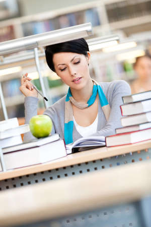 Woman with green apple surrounded with piles of books reads sitting at the table at the library....