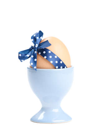 Colored easter egg with blue polka-dotted ribbon in blue egg cup, isolated on white. Traditional symbol of the feastの写真素材