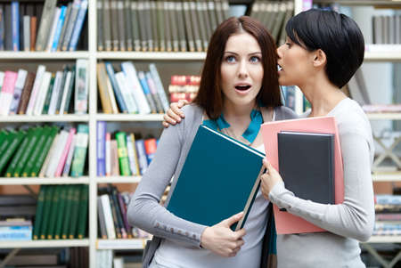 Two girlfriends keeping books whisper at the library. Friendship