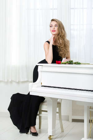 Full-length portrait of woman in black dress standing near the piano with red rose on it. Concept of music and artsの写真素材