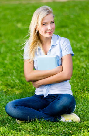 Female student holding book is ready to study. She sits on the green grass in the summer parkの写真素材