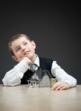 Portrait of little boy with home model and pile of coins on grey background. Concept of real estate and investmentの写真素材