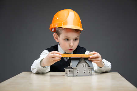 Portrait of little boy in headpiece with home model and ruler on grey background. Concept of real estate and engineeringの写真素材