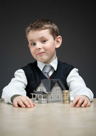 Portrait of little boy with house model and pile of coins on grey background. Concept of real estate...