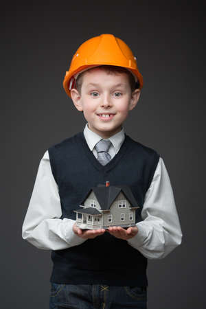Portrait of boy in hard hat keeping house model on grey backgroundの写真素材