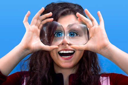 Smiling girl  holding two magnifying glasses up to eyes in a blue background
