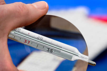 Medical mercury thermometer showing elevated body temperature in mans hand with magnifying glass