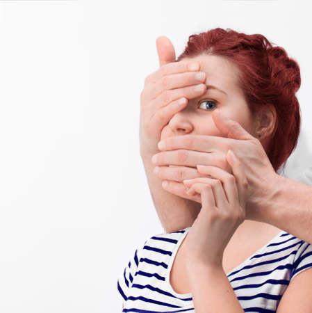 Man's hands covering caucasian redhair woman's mouth and one eye in a white backgroundの写真素材