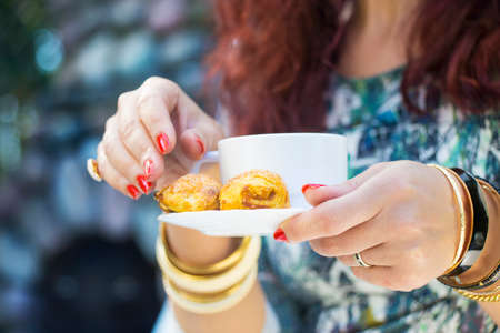 Woman's hands with golden bracelets, rings, cup of coffee and a snack on a plate