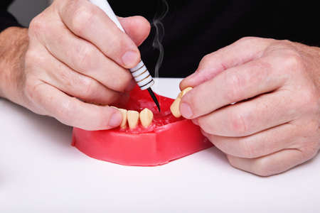 Close up of menâ€™s hands working with electric waxer on a red wax dental model on a white...