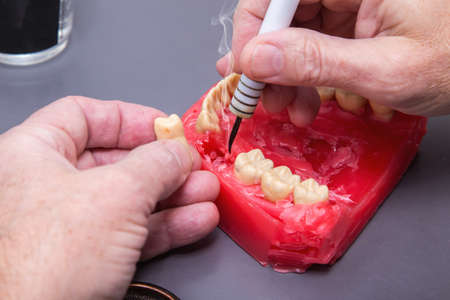 Close up of menâs hands working with electric waxer on a red wax dental model on a white tableの写真素材