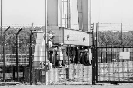 Bridge crane mechanisms behind barbed metal fence. Black white.