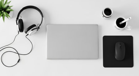 Clean, minimalist workspace featuring a closed laptop, headphones, coffee cups, and a mouse on a pad, all arranged on a bright white desk surface.の素材