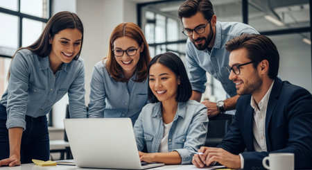 A diverse team of five professionals collaborates around a laptop in a bright, modern office, sharing ideas and smiling, showcasing teamwork and a positive work environment.の素材