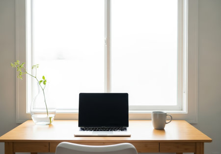 A bright, minimalist home office setup featuring a laptop, coffee mug, and a vase with greenery, set against a large window providing natural light.の素材