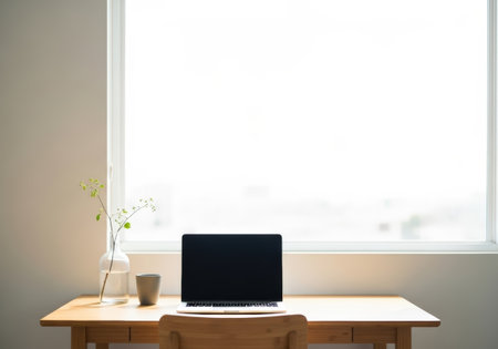 A minimalist workspace featuring a laptop, vase with greenery, and cup on a wooden desk, bathed in bright light from a large window, creating a serene and focused home office environment.の素材