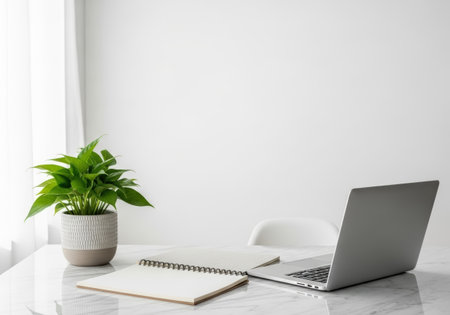 Clean, bright workspace featuring a laptop, open notebook, and a vibrant green potted plant on a marble desk against a white wall. Minimalist and modern aesthetic.の素材