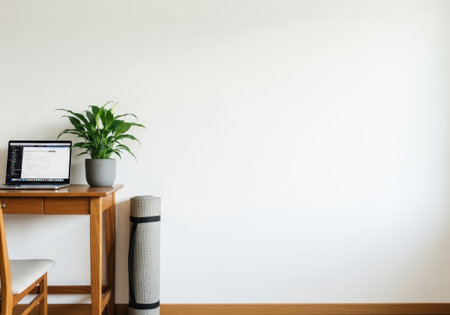 Clean, bright home office setup featuring a laptop, potted plant, and rolled yoga mat against a white wall, promoting wellness and productivity.の素材