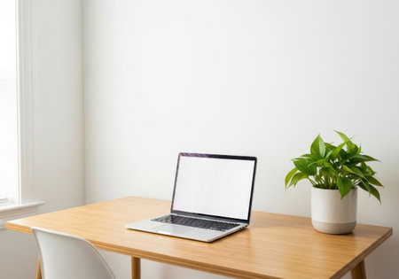 Clean, bright workspace featuring a laptop with a blank screen, a potted plant, and a wooden desk against a white wall, bathed in natural light.の素材