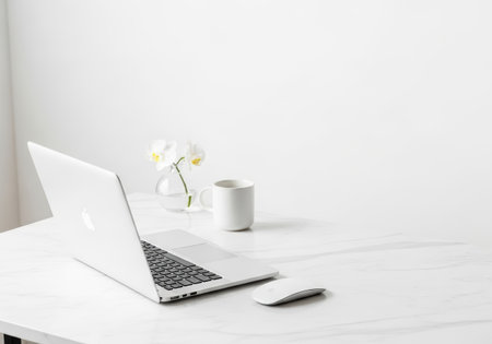 A clean, minimalist workspace featuring a silver laptop, white coffee mug, and a small vase with an orchid, all set on a white marble desk against a bright white background.の素材