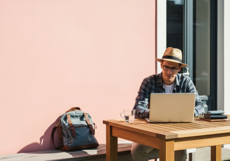 Man in hat and glasses works on laptop at wooden table outdoors. Backpack, water, and wallets nearby. Pink wall backdrop, bright daylight, casual workspace.の素材
