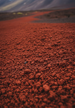 Close-up of red volcanic soil, resembling a Martian landscape, with distant mountains under a cloudy sky. The texture is granular, creating a unique and otherworldly scene.の素材