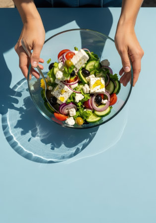 Hands toss a vibrant Greek salad in a clear glass bowl, featuring feta, tomatoes, olives, and red onion, set against a light blue backdrop with striking shadows.の素材
