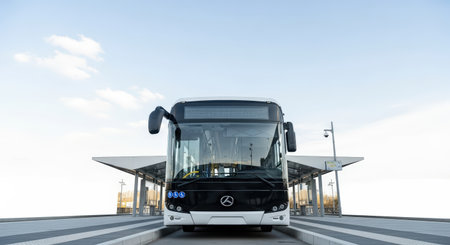 Front view of a modern Mercedes-Benz bus parked at a clean, minimalist bus station under a bright blue sky with scattered clouds. Focus on design and transportation.の素材