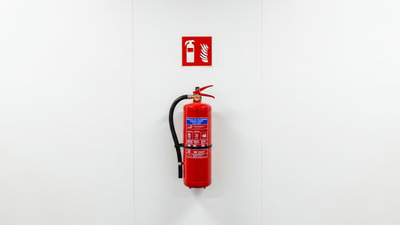 Bright red fire extinguisher with a black hose securely mounted on a pristine white wall, positioned beneath a clear safety sign Essential equipment for fire safetyの素材