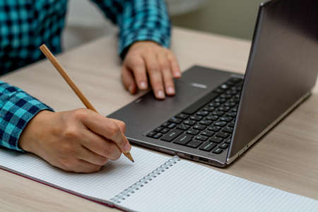 A woman in a plaid shirt writes with a pencil in a notebook and looks at the computer. The right...