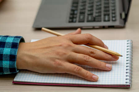 Female hand on blank notebook with brown pencil next to laptop. Horizontal view.
