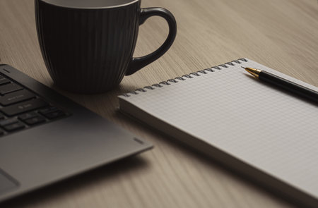 Close-up black pencil on a notebook next to a laptop and cup of coffee on a beige wood table. Horizontal view. Pencil tip in focus.の写真素材