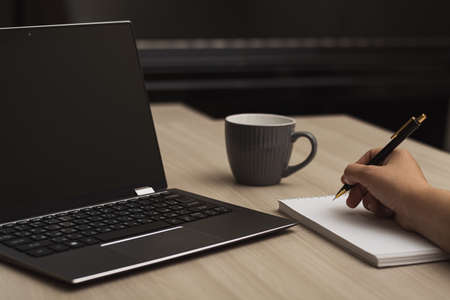 Close-up woman's hand holding a pencil and writing some information in a notebook next to a laptop...