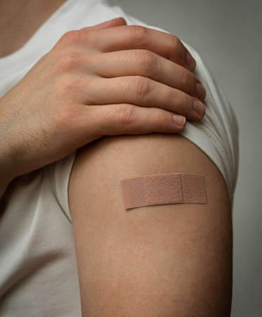 Close-up vaccinated man holds his shoulder. Adhesive plaster in focus. No face. White T-shirt. Gray...