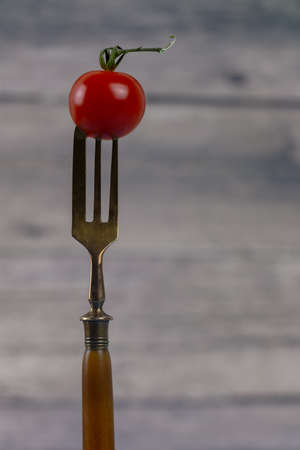 little red tomato skewered on a antique fork with three fork tines isolated on blurred wooden backgroundの写真素材
