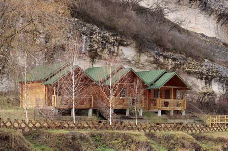 Three wooden houses stand in front of a snowy mountain on green grass.の写真素材