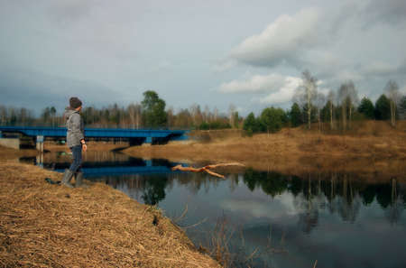spring river and reflections of trees and clouds in the water, a boy on the shore throws a branch into the waterの写真素材