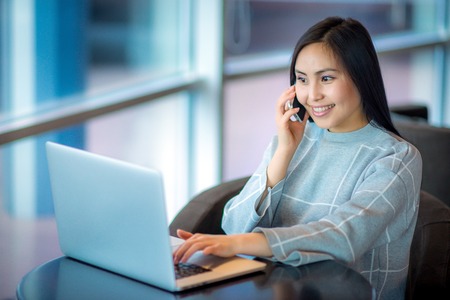 Young beautiful asian woman using laptop and talking on the phoneの写真素材
