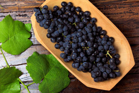bunches of fresh grapes with branches and leaves, inside wooden bowl, on rustic wooden tableの写真素材