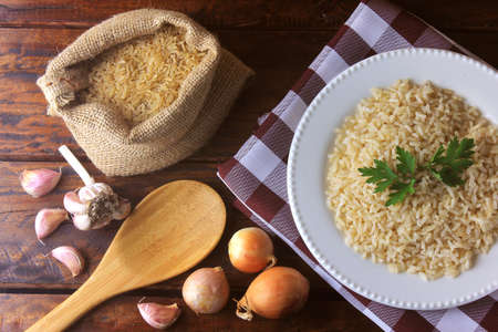 brown rice in the rustic bag next to ceramic dish and ingredients for cooking in the kitchen on rustic wooden table. Top viewの写真素材