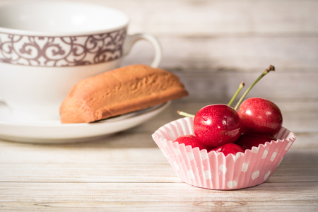 Studio shot of a coffee cup with a small cake and a cupcake liner with cherries, on wood background. Shallow depth of field.の写真素材