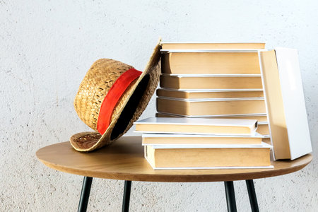 Books stacked next to a straw hat with a red ribbon, on a wooden table against a white wall. Copy...