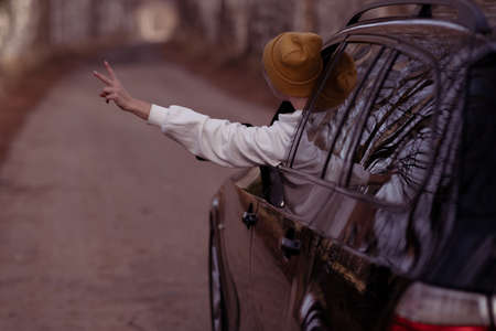 A girl in a tracksuit and a yellow cap shows her hand from the window of her black car. vacation and...