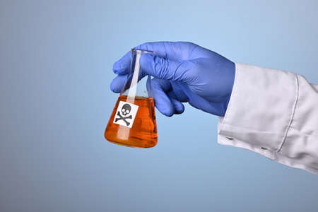 Doctor hand shows toxic orange fluid in a chemical flask. Medic hand wearing a blue nitrile glove holds glass retort with orange liquid poison. Scientific research to distinct death waste or remedy.の写真素材