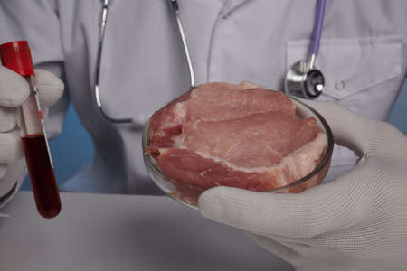 Blood test for artificial meat. Doctor holds vacuum blood test-tube and genetically grown meat in petri dish glass. Food control labs. Fake food production of GMO meat test-tube grown meat for vegans. Synthetic GMO food production and testing. How to speed up meat miocytes growth inside glass petri dish. Pork is growing in chemical glassware.の写真素材