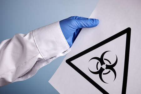 Doctor hand holds biohazard symbol printed on paper page. Labs photo with blue background. Bio-hazard sign inside black triangle on paper sheet. Scientist hand in blue nitrile glove. Danger symbol.の写真素材