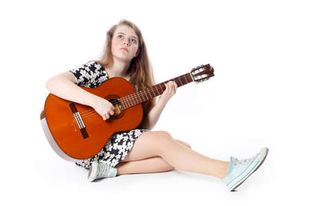 smiling teenage girl in dress plays the guitar sitting on the floor in studio with white backgroundの写真素材